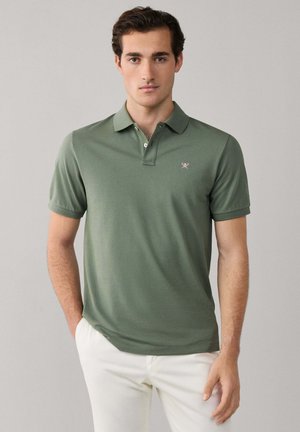 Jeune homme portant un polo vert avec un petit logo sur la poitrine et un pantalon blanc, se tenant devant un fond gris clair uni.