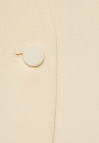 Tela beige con una textura suave que presenta un botón redondo y texturizado en un lado y un sutil detalle de costura.