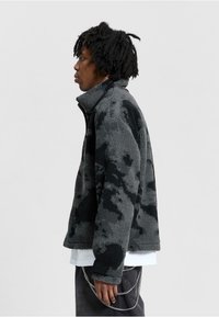 Giacca in pile con motivo camouflage nero e grigio, caratterizzata da un alto colletto, chiusura con zip e tessuto testurizzato. Mostrata su un modello.