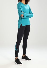 Türkises Sportoberteil mit langen Ärmeln und seitlichen Mesh-Einsätzen, kombiniert mit schwarzen Leggings mit türkisfarbenen Akzenten und einer figurbetonten Passform. Schwarze Sportschuhe.