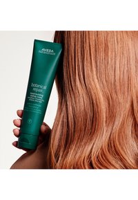 Aveda Botanical Repair bonding stylingkräm i ett mörkgrönt rör, hålls bredvid rakt, glänsande, rödbrunt hår.