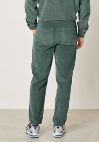 Zelene žametne hlače z teksturiranim koncem, z dvema zadnjima žepoma, kroj v obliki prilagojene prileganje, v kombinaciji s srebrnimi atletskimi čevlji.