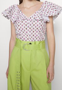 Blusa bianca con stampa floreale rosa e maniche a volant, abbinata a pantaloni verdi chiari a vita alta con una cintura larga e borchie decorative.