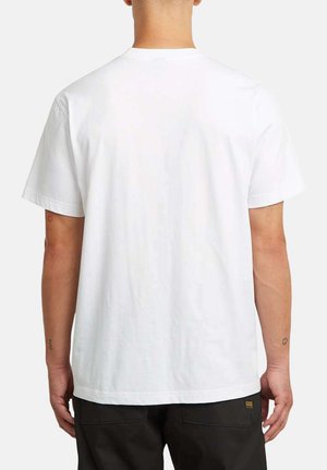 Personne portant un t-shirt blanc uni à manches courtes et un pantalon noir, vue de dos sur un fond clair uni.