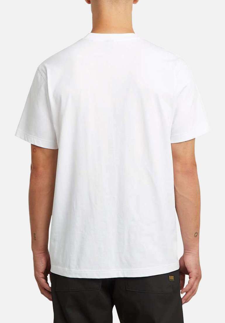 Persona vistiendo una camiseta blanca de manga corta y pantalones negros, mostrada de espaldas contra un fondo claro y liso.