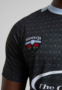 Juodas trumpas rankovėmis džersis su raštuotu dizainu ir pleistru su užrašu „BROOKLYN“ bei rožių grafika. Matomi šviesiai mėlyni apykaklės akcentai.