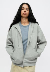 Ανοιχτό γκρι φούτερ με φερμουάρ και κουκούλα, μπροστινές τσέπες και κορδόνια, φορεμένο πάνω από μπλε μπλούζα, το οποίο εμφανίζεται σε απλό φόντο.