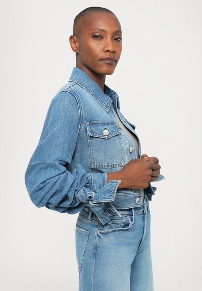 Person with short hair wearing a light grey top, blue denim jacket, and blue jeans, standing sideways against a plain white background.