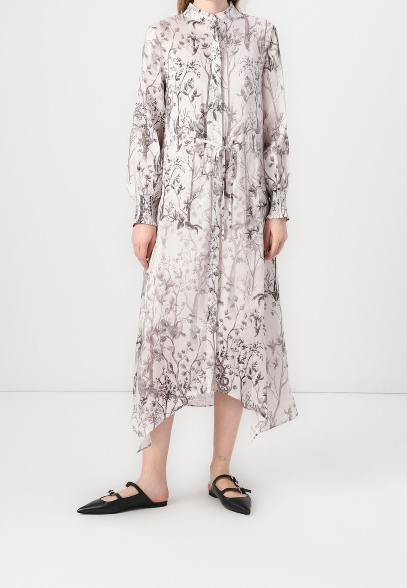 Femme portant une robe rose clair à manches longues boutonnée avec des motifs floraux gris et des chaussures plates noires pointues avec des brides à la cheville.