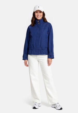 Woman wearing a navy jacket, white pants, white sneakers, and a white cap, standing and smiling against a plain background.