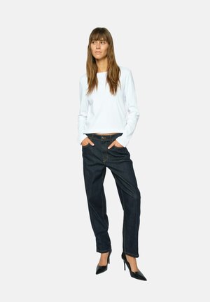 Donna con lunghi capelli castani che indossa una maglia bianca a maniche lunghe, jeans scuri, tacchi neri, in piedi con le mani in tasca.