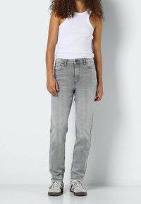 Jeans de mezclilla gris con corte recto, con un clásico diseño de cinco bolsillos y un desgaste sutil. Combinados con una camiseta sin mangas blanca y zapatillas.