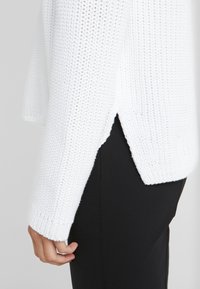 Biały sweter dziany z bocznymi rozcięciami, długimi rękawami i ściągaczami na mankietach. Połączony z czarnymi spodniami, podkreślając teksturę i wzór.