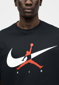 Μαύρο φούτερ με μεγάλο λευκό λογότυπο Nike Swoosh και κόκκινο λογότυπο Jumpman με την επιγραφή "AIR" από κάτω. Υφασμάτι νο από βαμβάκι, σχεδιασμός με στρογγυλή λαιμόκοψη.