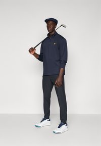 Golf outfit featuring a navy long-sleeve polo with yellow logo, dark trousers, and white athletic shoes with blue accents. Golf club held on shoulder.