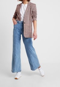 Rutig kavaj i röd, beige och svart; matchad med högmidjade, vida ljusblå jeans och vita sneakers med tan-färgad sula.