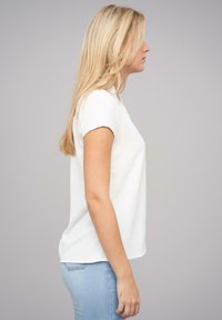 Top bianco a maniche corte con finitura testurizzata, scollatura rotonda e vestibilità morbida. Abbinato a jeans azzurri, presentato in vista laterale.
