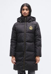 Μαύρο μπουφάν puffer με κουκούλα, που κλείνει με φερμουάρ, με λεπτομέρεια χρυσού εμβλήματος και καπιτονέ υφή. Μακριά μανίκια και μήκος έως τη μέση.