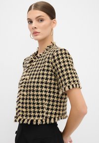 Kortärmad jacka i svart och beige hundtandsmönster med fransdetaljer och guldfärgade knappar, som har en kortad design och texturerad tyg.