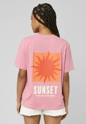 Femme aux cheveux tressés portant un t-shirt rose avec un motif de soleil orange et le texte « SUNSET Glow now, worry later », assorti à un short blanc.