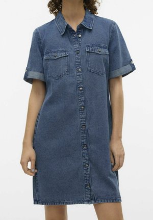 Femme portant une robe en denim à manches courtes, boutonnée, avec deux poches poitrine et des manches retroussées, sur un fond uni.