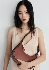 Bruine leren handtas met een gebogen vorm en contrasterende stiksels. Draagbaar als crossbody met een verstelbare schouderband. Glad textuur.