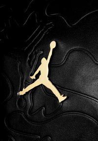 Fekete textúrájú felület arany Jumpman logó ikonral, kiemelkedő mintázattal, sima és matt felületek kontrasztjával.