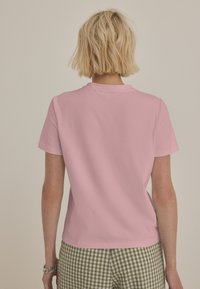 Rožnata majica s kratkimi rokavi iz bombaža, z okroglim izrezom in gladko teksturo, prikazana od zadaj, kombinirana s karirastimi zelenimi kratkimi hlačami.