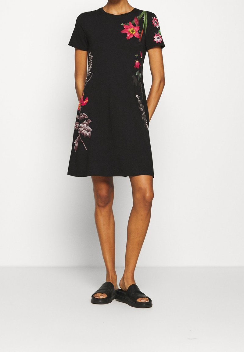 Schwarzes kurzärmeliges Kleid mit floralem Stickerei in Rosa und Grün, mit lockerer Passform und seitlichen Taschen. Getragen mit schwarzen Rutschen-Sandalen.