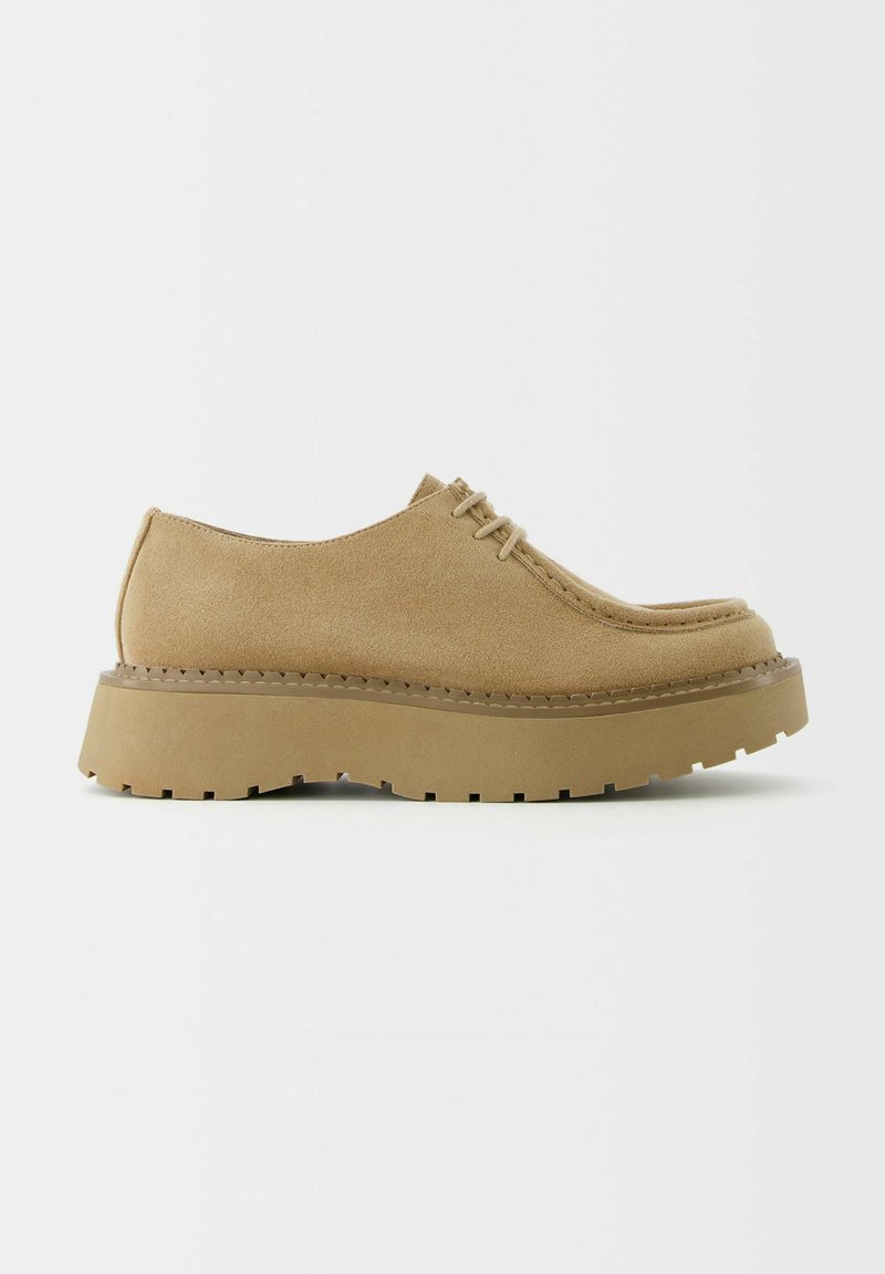 Zapatos de gamuza color tostado con cordones, punta redondeada, costuras prominentes y una suela de plataforma gruesa y texturizada con borde aserrado.