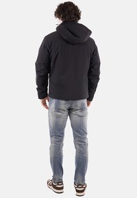 Uomo con capelli ricci che indossa una giacca nera con cappuccio, jeans blu sbiaditi e sneakers Nike marroni, in piedi di spalle.