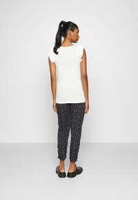 Top branco sem mangas com ombros franzidos, combinado com calças pretas com bolinhas e sapatos pretos sem fecho. O tecido parece suave e leve.
