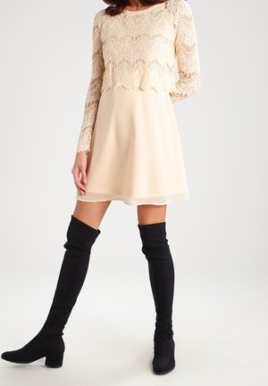 Frau trägt ein cremefarbenes Spitzenoberteil mit langen Ärmeln und einen passenden Rock, dazu schwarze kniehohe Stiefel mit Absatz, steht vor einem einfachen Hintergrund.