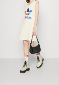 Beżowa sukienka bez rękawów Adidas z kolorowym logo, zestawiona z pasiastymi skarpetkami i białymi butami wojskowymi. Trzyma małą czarną torbę.