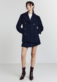 Marineblauer Peacoat mit doppelt geknöpften Knöpfen, großem Kragen und Seitentaschen, kombiniert mit einem plissierten marineblauen Rock und glänzenden burgunderfarbenen Stiefeletten.