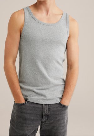 Homme portant un débardeur gris clair sans manches avec les mains dans les poches d’un jean gris foncé sur un fond uni.