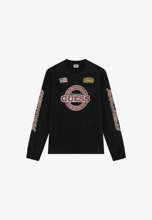 Schwarzes Langarmshirt mit "GUESS"-Logo und Patches, das einen strukturierten Druck sowie das Wort "International" in Rot entlang des Ärmels aufweist.