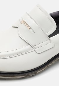 Białe skórzane mokasyny z przeszyciami, paskiem na górze, beżową metką oraz czarną podeszwą z wzorkiem.