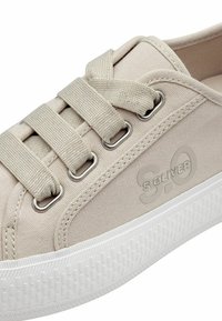 Zapatilla de lona beige con cordones de tela, ojetes de metal y una suela de goma blanca. Presenta un logo en relieve en el lateral y un diseño texturizado en la suela.