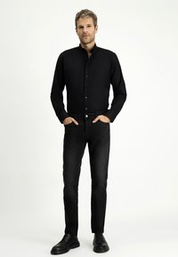 Camicia nera con colletto abbinata a jeans neri slim-fit e scarpe nere. Il modello si trova in piedi con le mani in tasca su uno sfondo neutro.