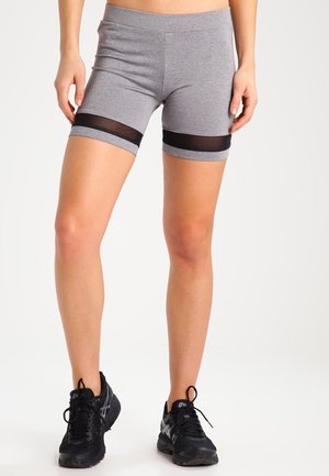 Vrouw die grijze sportshorts met zwarte meshstrepen en zwarte hardloopschoenen draagt, staand tegen een effen achtergrond.