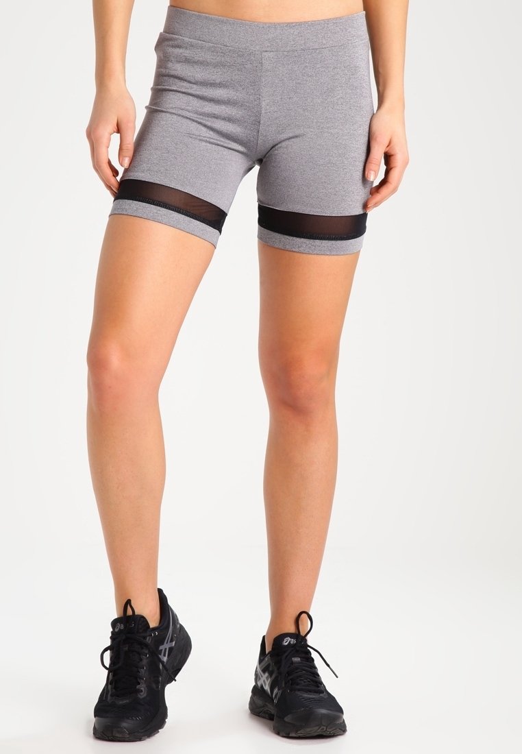 Femme portant un short de sport gris avec des bandes en mesh noir et des chaussures de course noires, debout devant un fond uni.