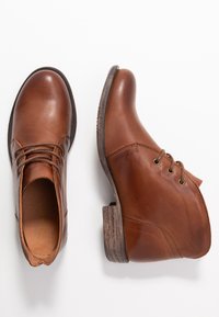 Stivali ankle in pelle marrone con punte rotonde, lacci davanti, tacco basso impilato e suole in gomma testurizzata su sfondo bianco.