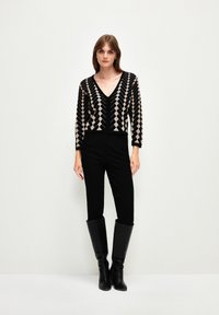 Pull court à motifs noir et beige avec un col en V et des manches longues, associé à un pantalon noir ajusté et des bottes hautes noires.