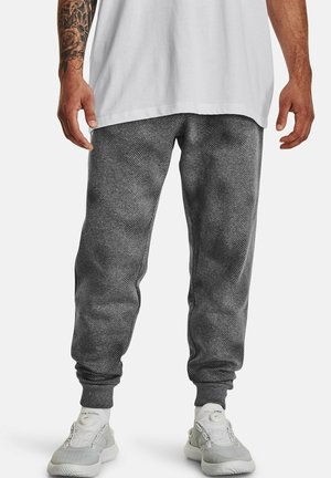 Man draagt donkergrijze textuur joggingbroek, witte sokken en grijze sportschoenen, met zichtbare getatoeëerde linker onderarm.