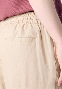 Beiga linnebyxor med elastisk midja och en bakficka. Designen har en diskret broderad detalj nära fickan.