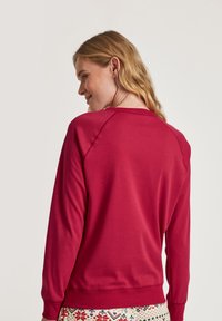 Red sweatshirt with a crew neck and long raglan sleeves, featuring smooth fabric and ribbed cuffs, viewed from the back.