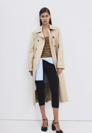 Ženska v bež trenč plašču čez rjavo črtasto majico, črne skrajšane hlače, svetlo modra srajca zavezana v pasu in črne čevlje z visokimi petami.