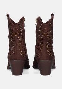 Botas de tobillo de ante marrón con tachuelas decorativas, puntera afilada y un tacón bajo de bloque. Presentan cremalleras laterales y recortes curvados en la parte superior.
