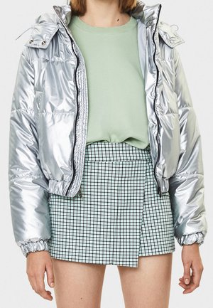 Osoba nosící lesklou stříbrnou péřovou bundu, světle zelený svetr a vysokým pasem kostkovanou minisukni s asymetrickým lemem.